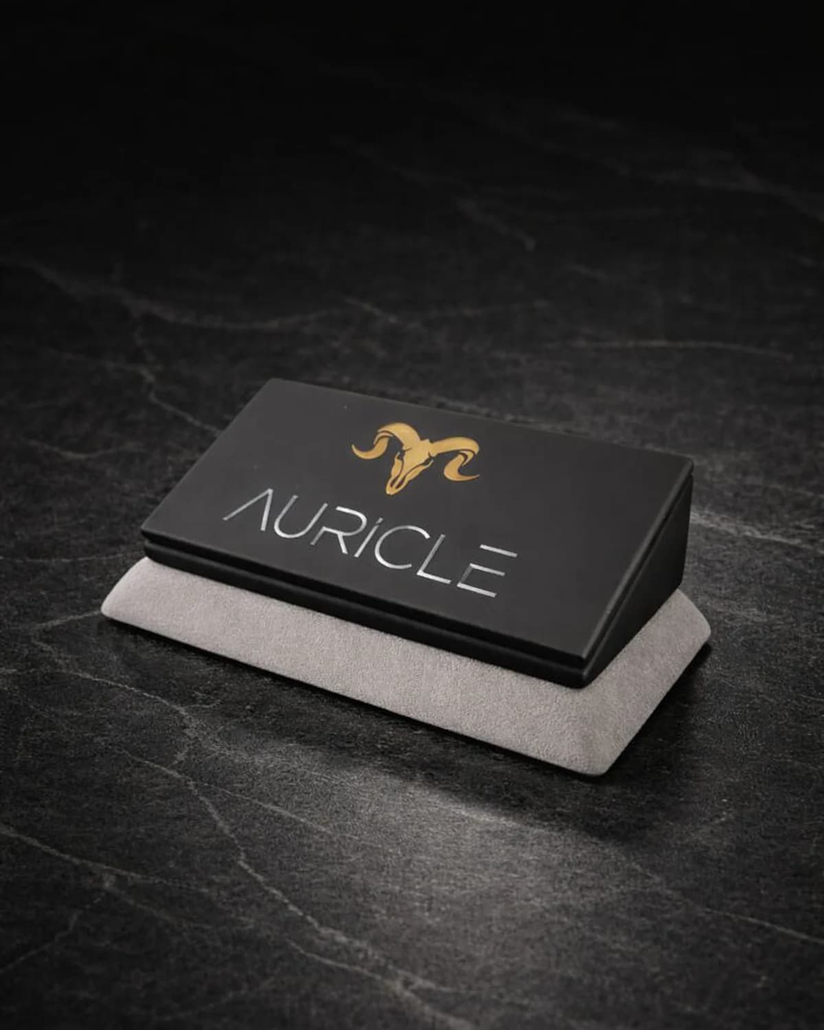 AURICLE jewellery branded nameplate stand. The stand has a light gray suede base with a black leather top. Sat on a dark worktop with moody lighting.